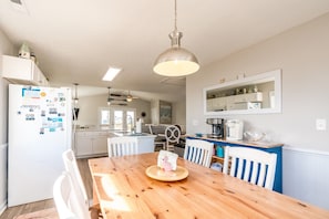 Une cuisine avec une table en bois, des chaises blanches, un réfrigérateur et un luminaire suspendu.