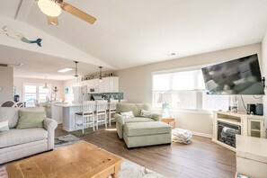 A modern living room with a sofa, ottoman, and coffee table. A kitchen with white cabinets and a dining area with white chairs is visible in the background. - Knot Too Shabby - 4 BR Home with Covered Porch and Ocean View (North Topsail Beach)