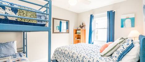 A room with bunk beds, a ceiling fan, a window with curtains, a bookshelf, and a bed with a floral bedspread.