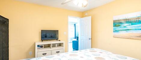 Un dormitorio con cama, ventilador de techo, televisor sobre un mueble, una puerta que da a otra habitación y un cuadro con una playa.