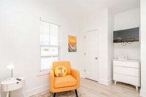 A room with a white wall, a window with blinds, a white side table with a lamp, an orange chair with a cushion, a white dresser, and a flat-screen TV mounted on the wall. - 613A Follow the Sun - Upscale 4 BR Home Surf City; Dogs welcome! (Surf City)