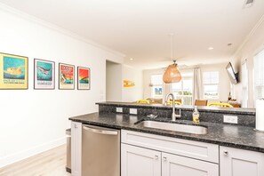A modern kitchen with a granite countertop, stainless steel sink, and a dishwash unit. There are framed posters on the wall and a view of the living area with a yellow chair and a table. - 613A Follow the Sun - Upscale 4 BR Home Surf City; Dogs welcome! (Surf City)