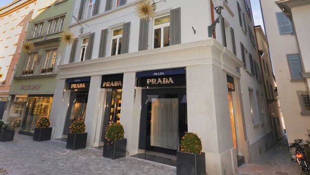 Un edificio de varios pisos con locales comerciales en la planta baja, incluyendo una tienda Prada. La arquitectura presenta un diseño clásico con techo plano y ventanas simétricas.