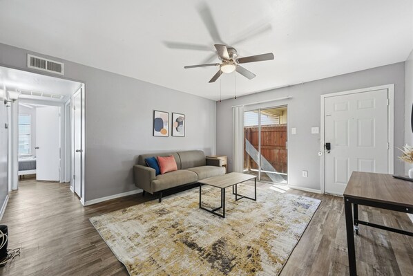 A living room with a sofa, coffee table, and a ceiling fan. There is a door leading to a balcony and a window with blinds.