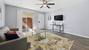 Ein Wohnzimmer mit grauem Sofa, Couchtisch, gemustertem Teppich, Deckenventilator, einer Glasschiebetür und einem Fernseher auf einem Standfuß.