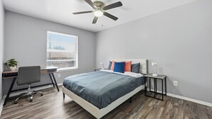 Ein Schlafzimmer mit Bett, Schreibtisch, Stuhl, Deckenventilator und Fenster mit Jalousien.