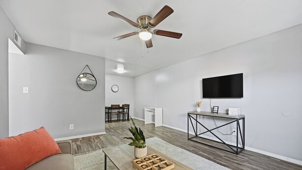 A modern living room with a ceiling fan, a coffee table with a plant, a TV on the wall, and a dining area in the background. - Denton pad with 2BR comfort (Denton)