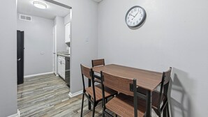 A dining area with a wooden table and chairs, a clock on the wall, and a kitchen in the background. - Denton pad with 2BR comfort (Denton)