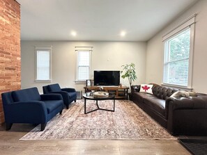 A living room with a brick wall, two blue sofas, a brown leather sofa, a coffee table, a TV, and a plant. - Trendy Home - Historic Walkerville Distillery District (Windsor)