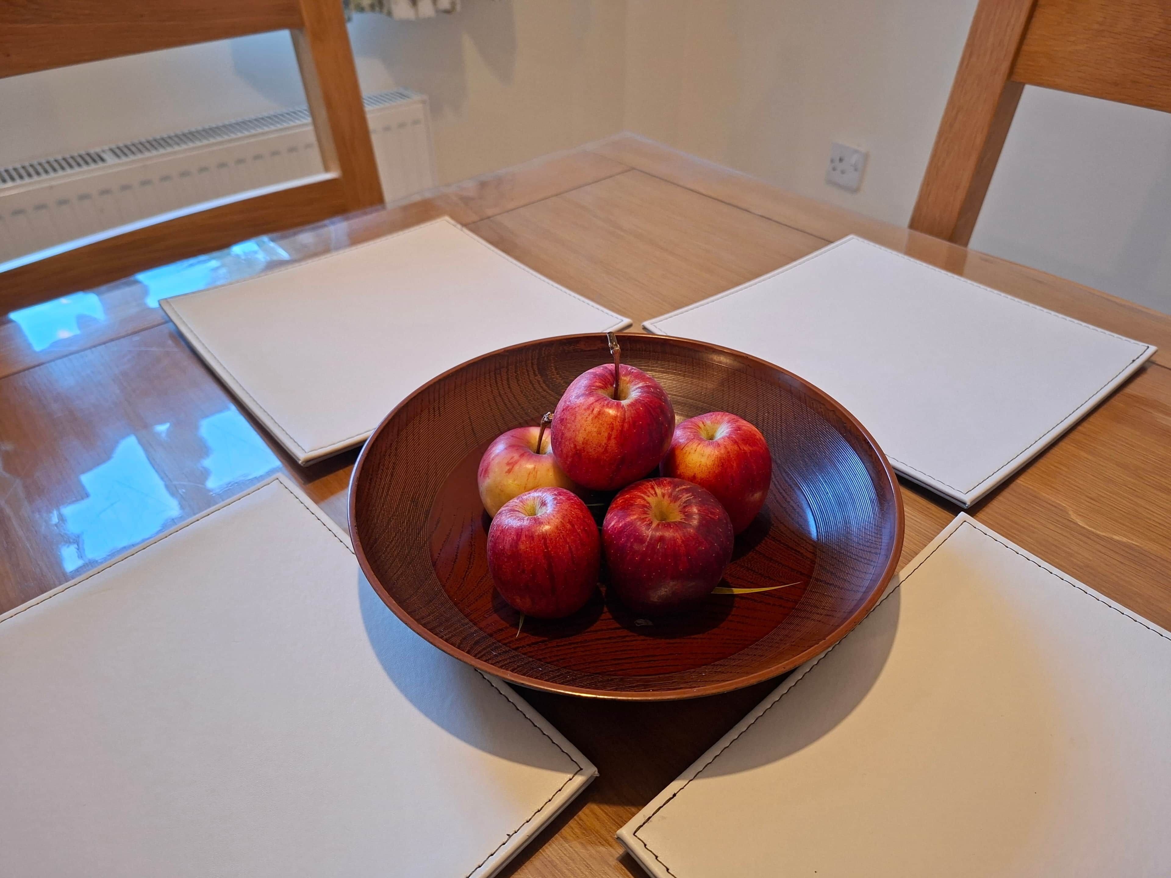 A wooden table with a brown bowl containing apples, surrounded by white placemats.