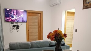 A living room with a grey sofa, a dining table with chairs, a vase with flowers, and a TV mounted on the wall. - Luxury and Peace Near Downtown Puntacana (Punta Cana)