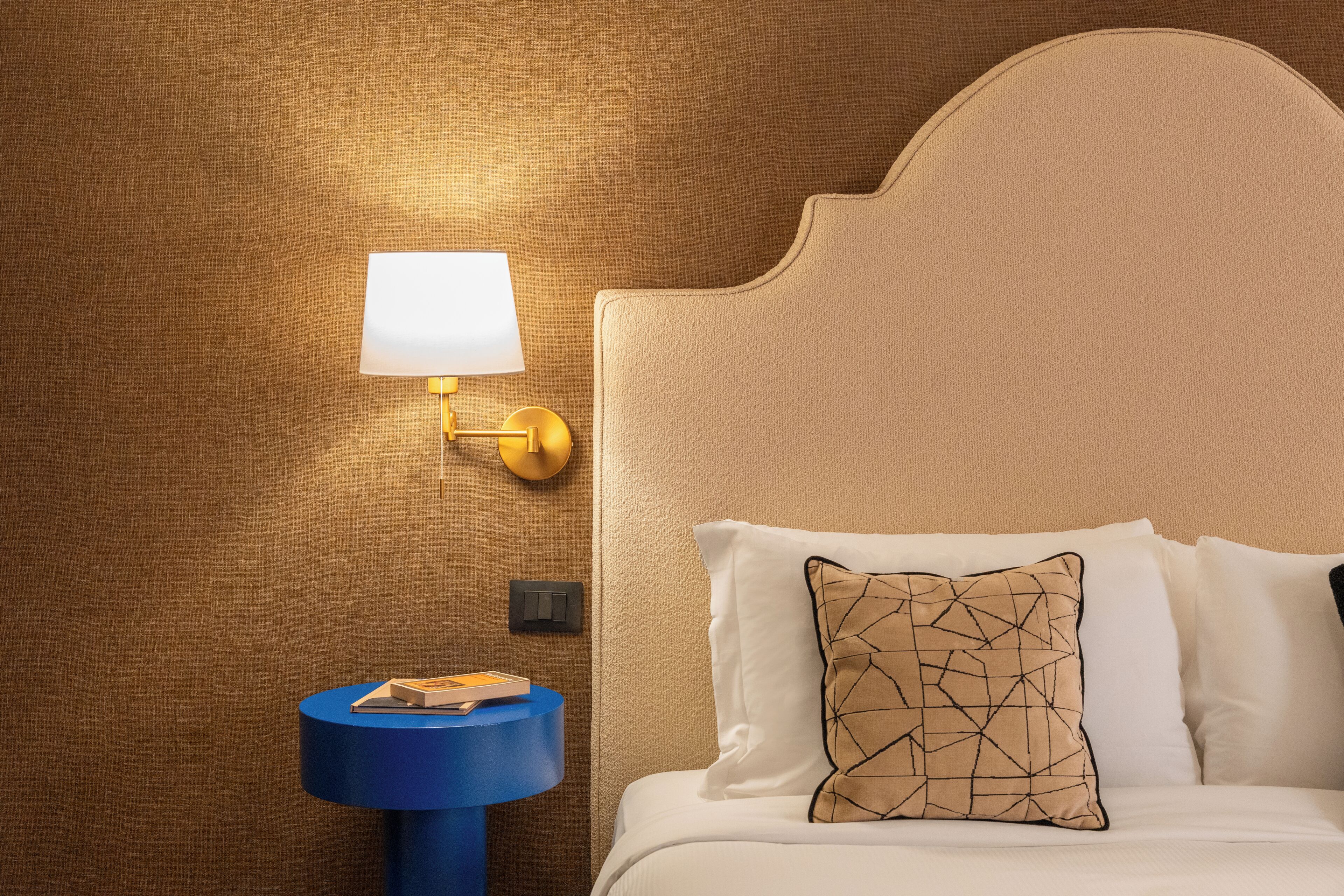 Une chambre d’hôtel avec un lit, une lampe de chevet, une petite table avec un livre et un interrupteur mural.