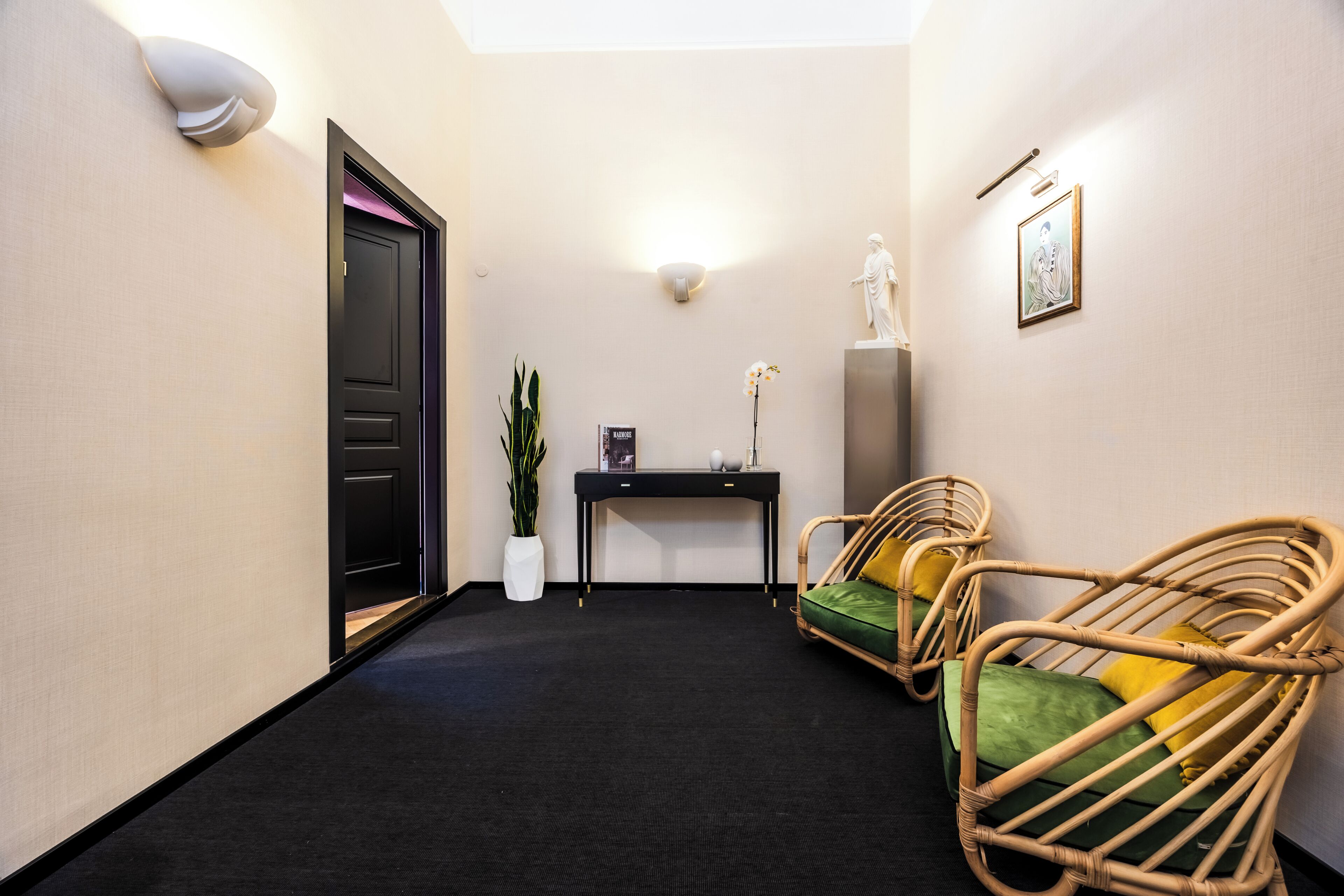 A hallway with a black carpet, a black door, a white wall-mounted light, a black console table with a vase and a book, and two wicker chairs with green cushions.