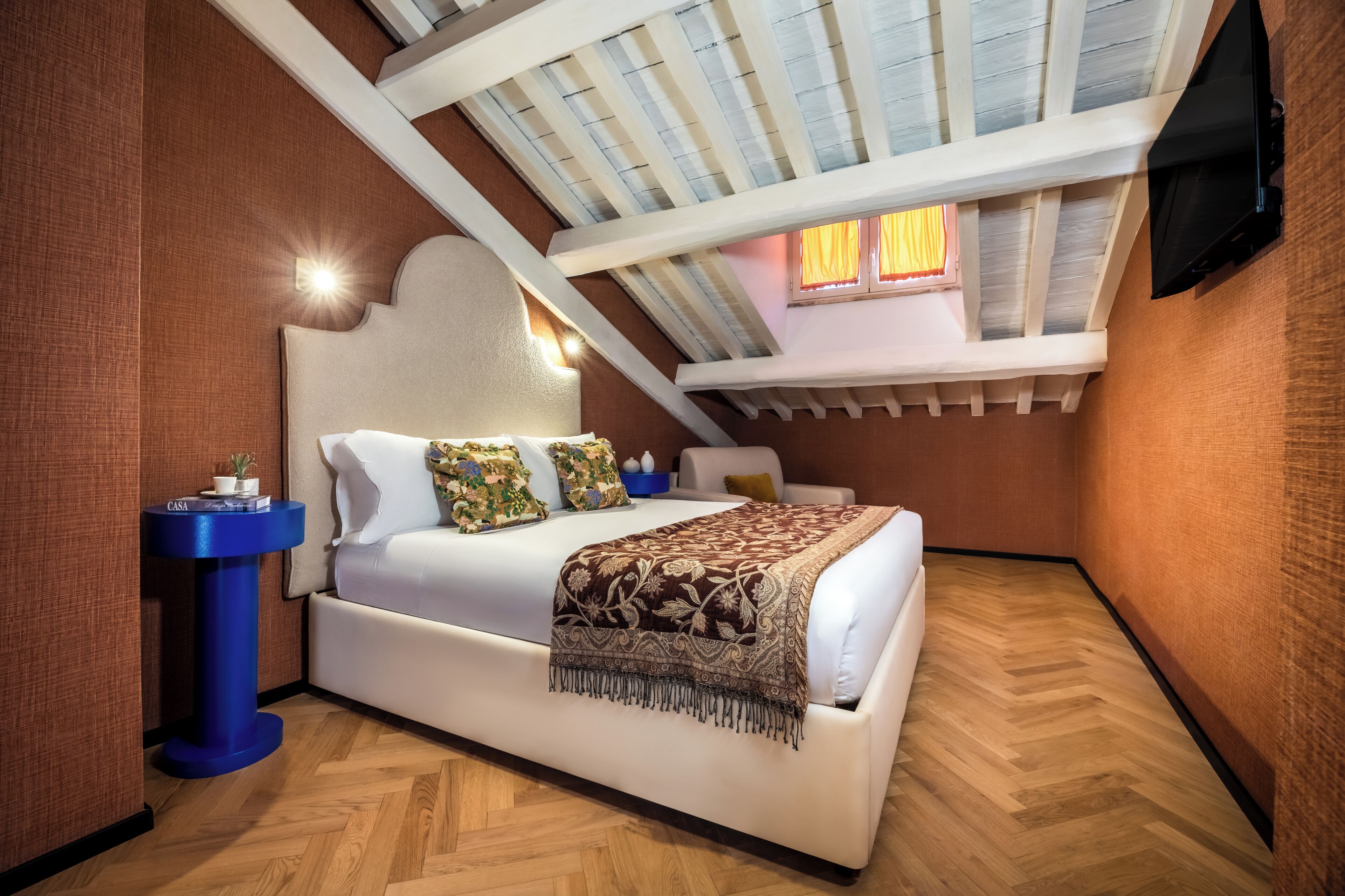 A bedroom with a sloped ceiling, a bed with a patterned blanket, a bedside table with a plant, and a window with curtains.