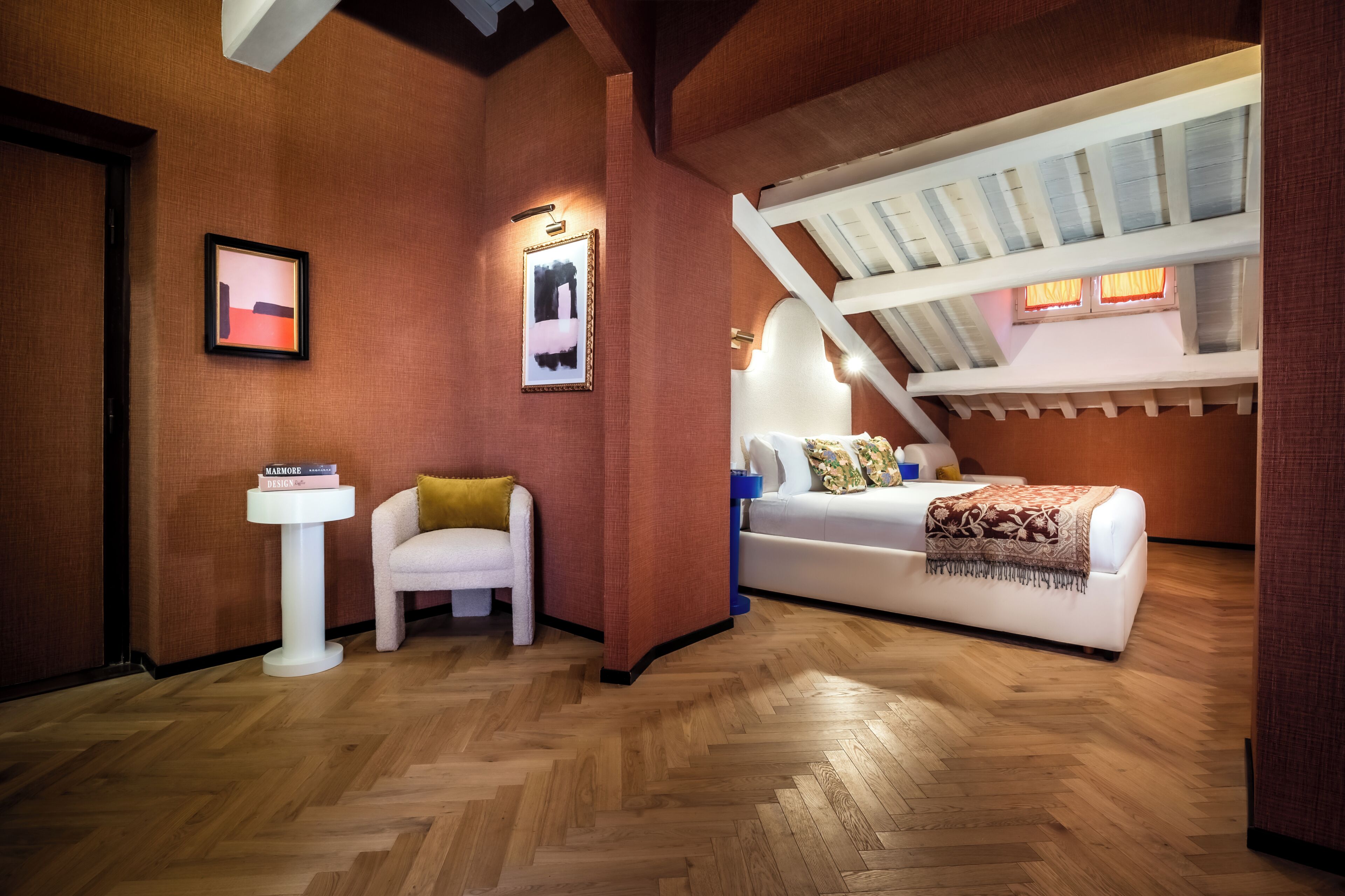 A room with a wooden herringbone floor, a white sofa with a patterned throw, a white chair, a small white pedestal table, and two framed pictures on the wall.