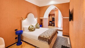 Une chambre d’hôtel avec un lit, une table de chevet bleue, un plaid à motifs et une vue sur la salle de bain à travers une arche.