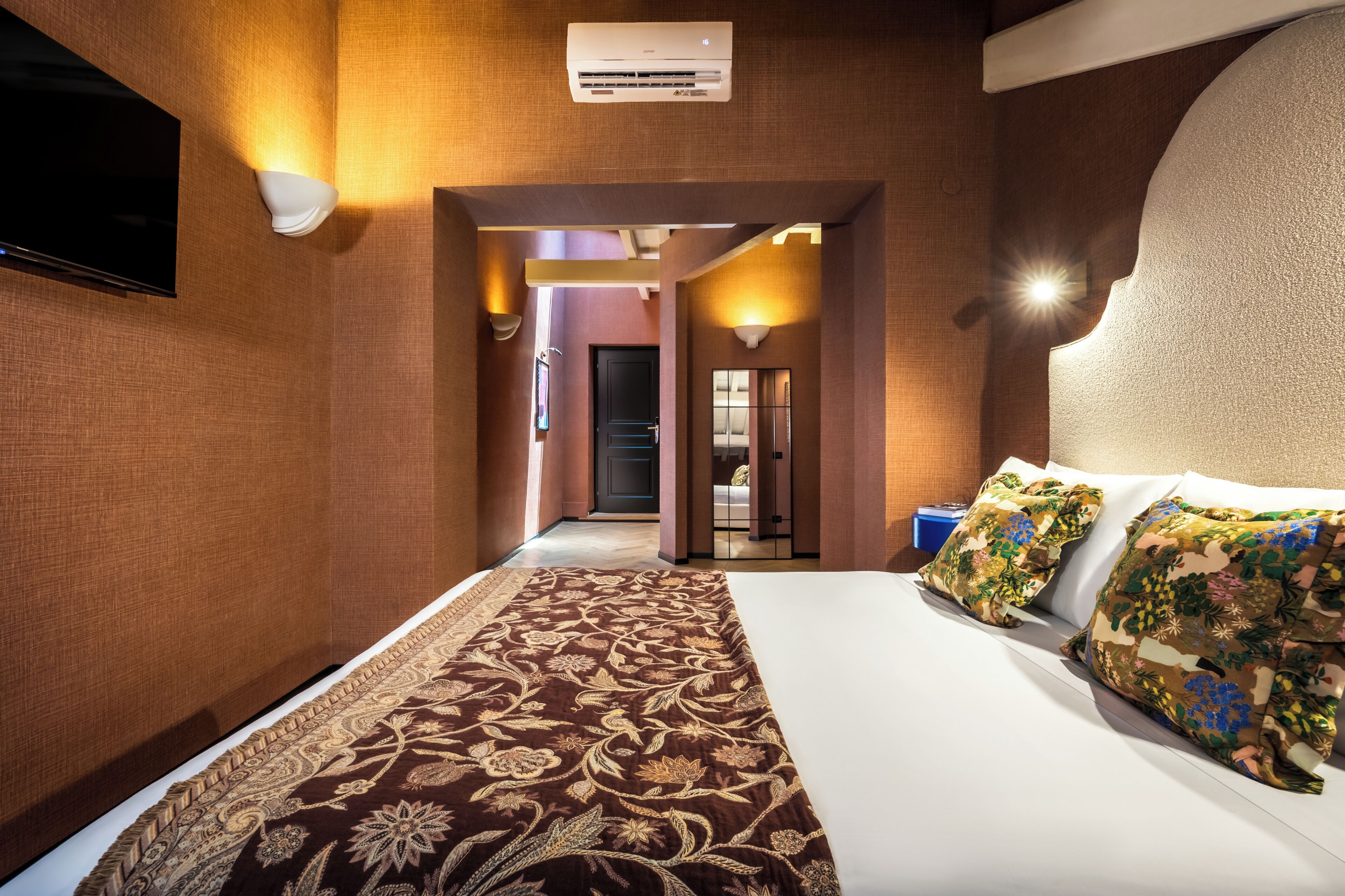 A hotel room with a large bed, a flat-screen TV, a patterned rug, and a hallway leading to another room.