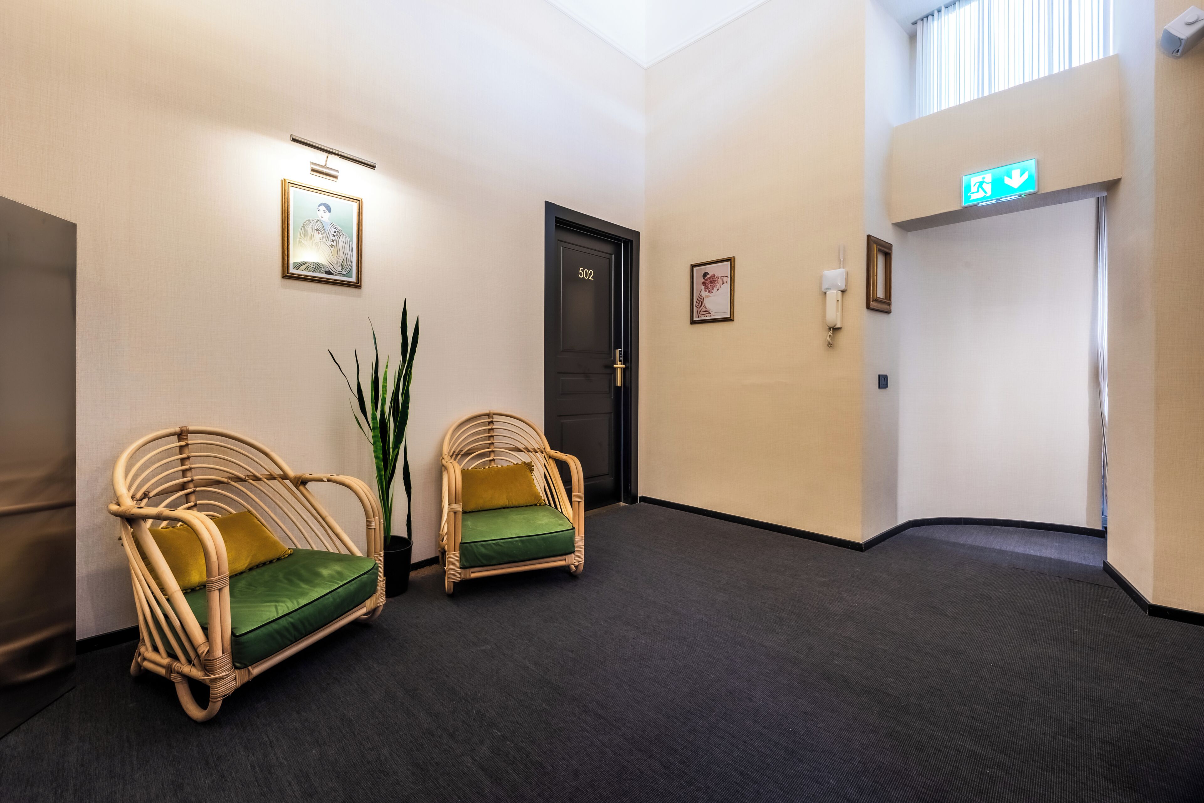 A hallway with two wicker chairs, a door labeled 502, and a green exit sign.