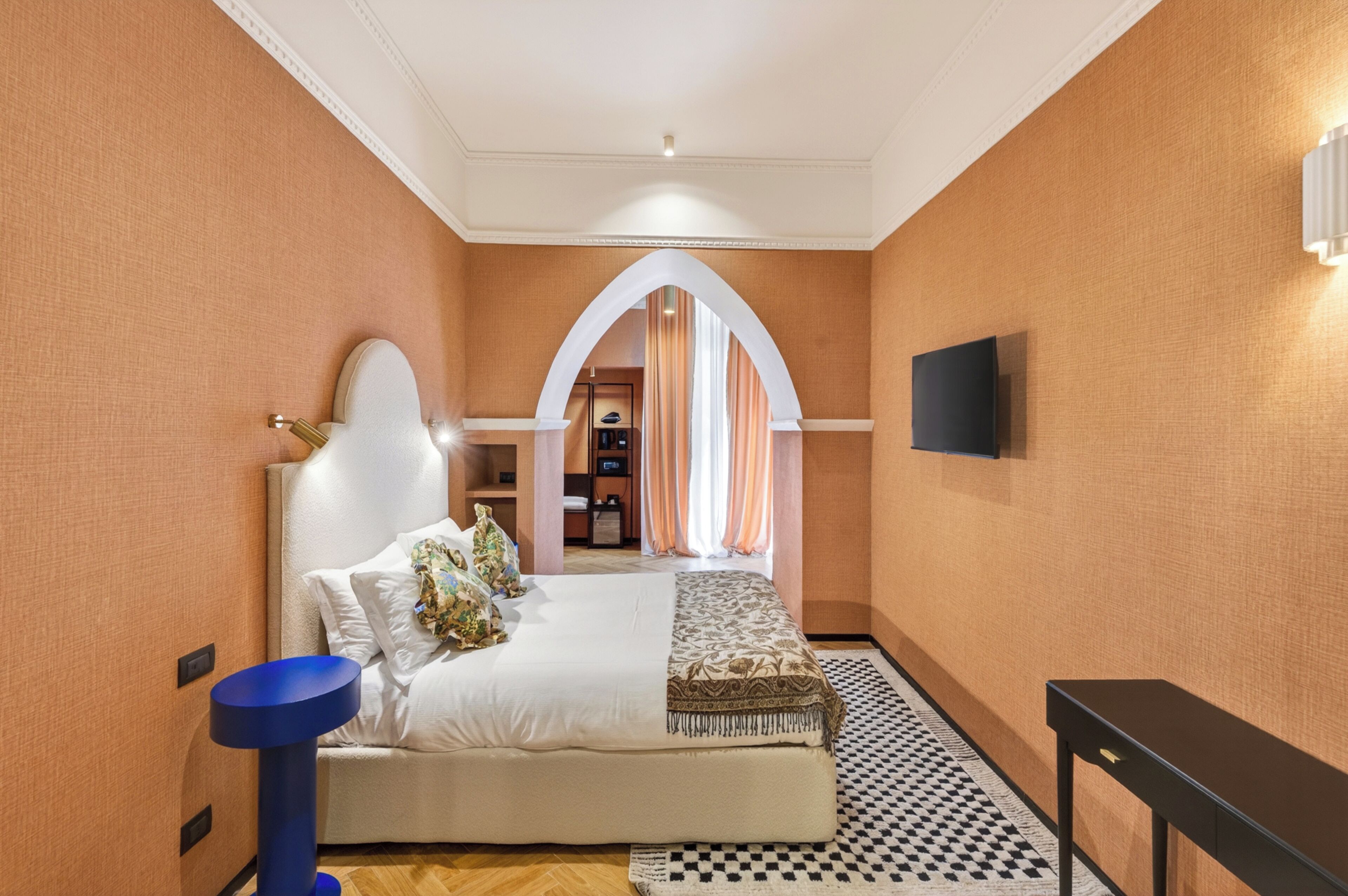 A hotel room with a bed, a blue side table, a black side table, a TV mounted on the wall, and a patterned floor.