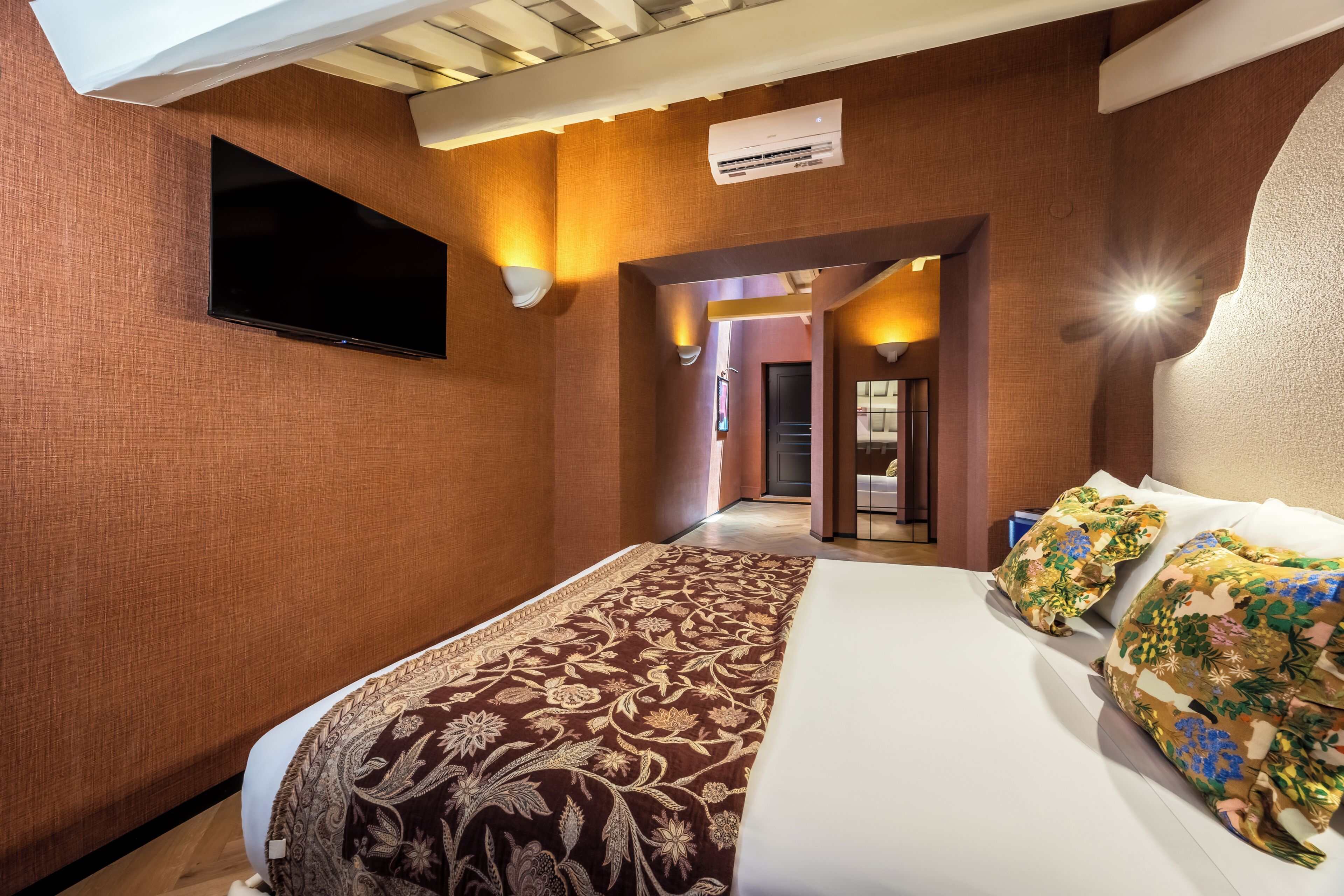 A hotel room with a large bed, a flat-screen TV, a patterned bedspread, and a hallway leading to another room.