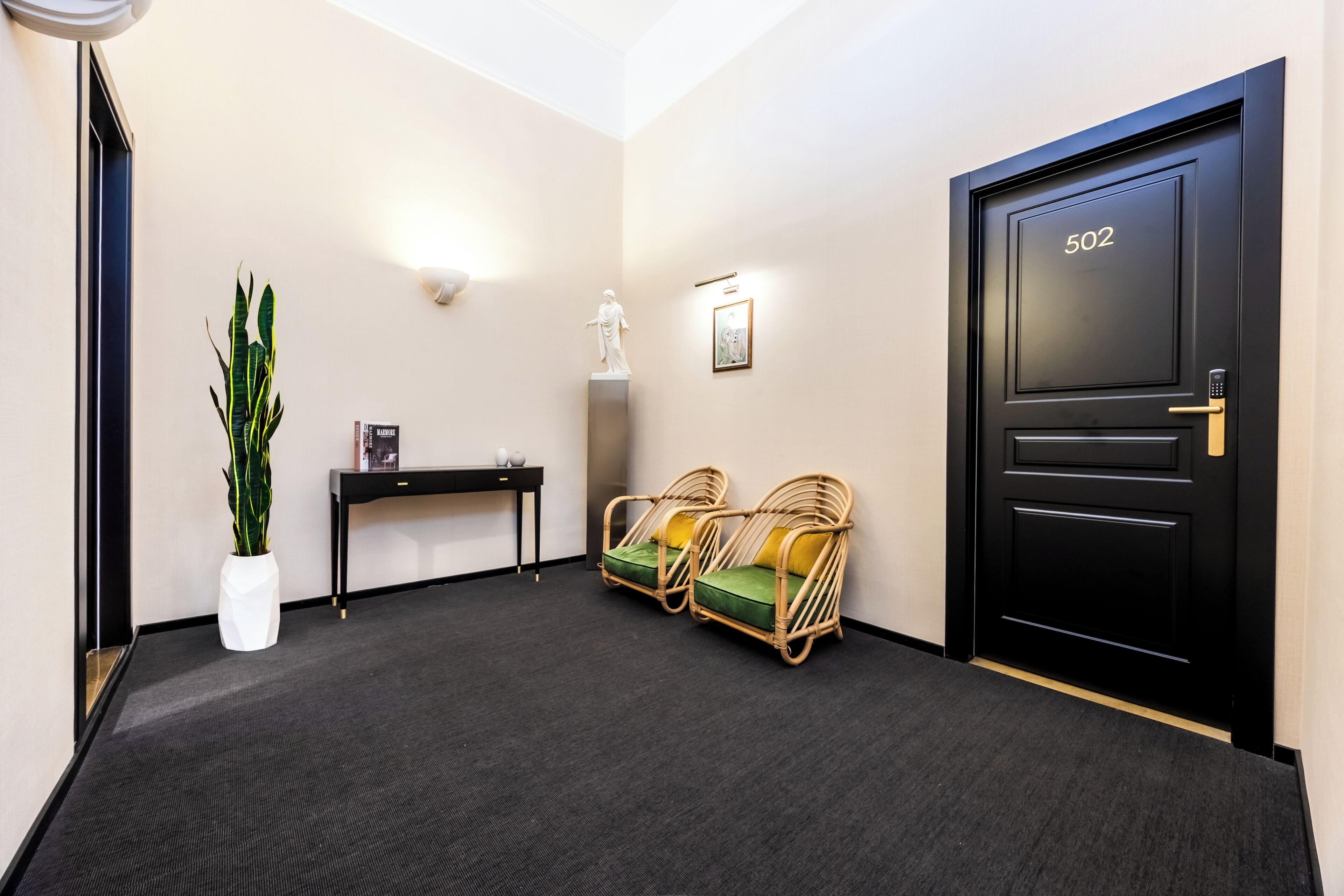 A hallway with a black door marked '502', a white statue, two wicker chairs with cushions, a small table with a lamp, and a potted plant.
