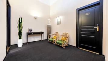 A hallway with a black door marked '502', a white statue, two wicker chairs with cushions, a small table with a lamp, and a potted plant.