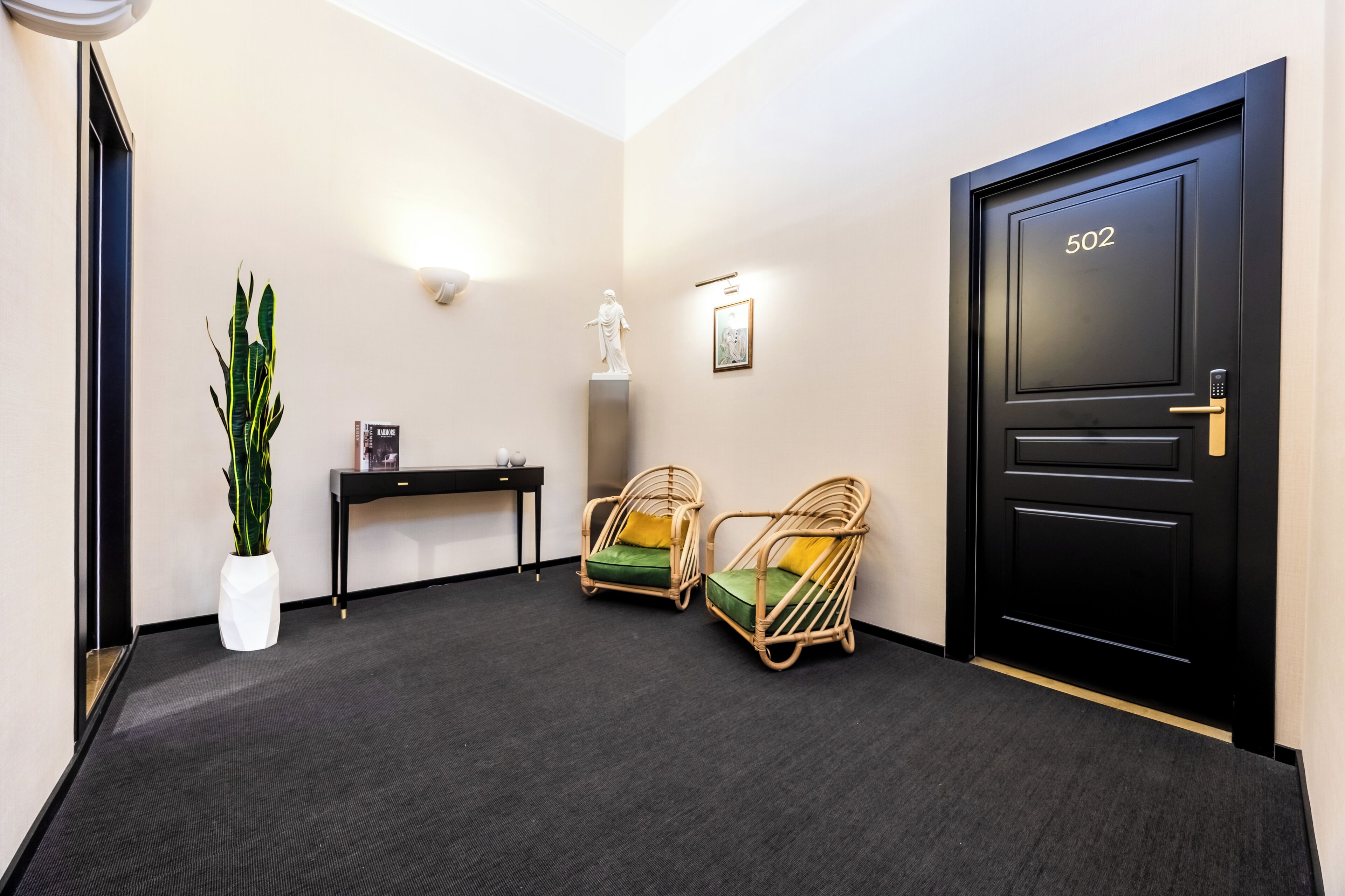 A hallway with a black door labeled '502,' a potted plant, two wicker chairs with green cushions, a small table with a lamp, and a framed picture on the wall.