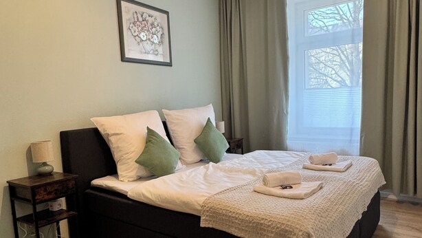 A bedroom with a bed, a nightstand, a framed picture, and a window with curtains. - Arminos Apartment (Chemnitz)