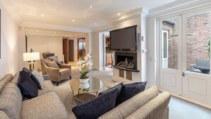 A modern living room with a sofa, armchair, and coffee table. There is a fireplace with a TV mounted above it. - Luxury 6BR Knightsbridge house w/ Parking (London)