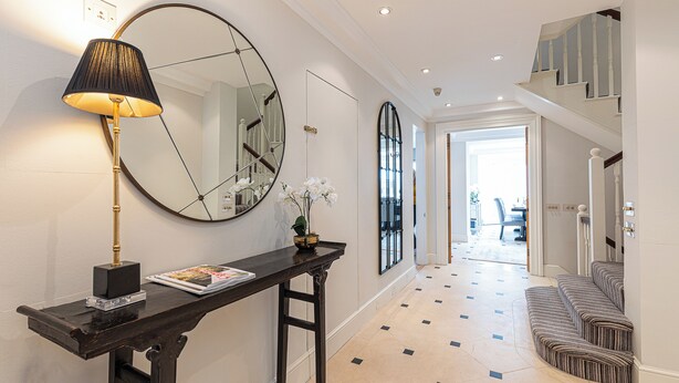 A hallway with a dark wooden console table, a large mirror, and a lamp. There is a staircase with a white railing leading to an upper level. - Luxury 6BR Knightsbridge house w/ Parking (London)