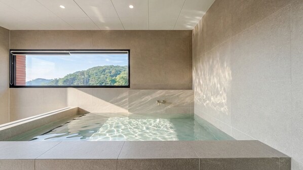Ein modernes Badezimmer mit einer großzügigen Badewanne, einem Fenster mit Blick auf einen bewaldeten Hügel und einem gefliesten Boden.