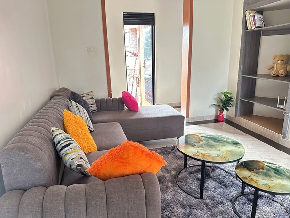 A modern living room with a grey sectional sofa, two glass coffee tables, and a bookshelf with books and a teddy bear.