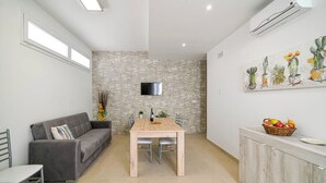 Un espacio habitable moderno con una pared de piedra, un televisor de pantalla plana, una mesa de comedor con sillas, un sofá gris y un mueble con fruta.