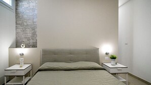 Un dormitorio con una cama, dos mesitas de noche, una mesita de noche con lámpara, un reloj en la pared y una planta en maceta.