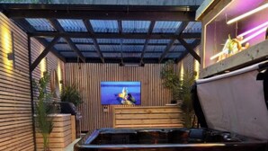 An outdoor patio area with a wooden bench, a hot tub, and a television screen. - Stylish City Centre Hideaway & Hot Tub (Galway)