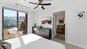 Un dormitorio moderno con una cama grande, un escritorio, una silla y un ventilador de techo. Hay un balcón con vistas a las montañas.