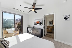 Ein modernes Schlafzimmer mit einem großen Bett, einem Schreibtisch, einem Stuhl und einem Deckenventilator. Ein Balkon bietet Blick auf die Berge.