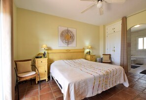 A hotel room with a bed, two bedside tables with lamps, a chair, a painting on the wall, and a bathroom visible through an archway. - Charming villa with private pool in Mijas Costa (Mijas)