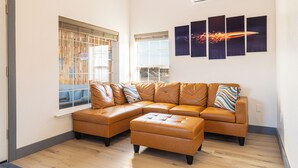 Een moderne woonkamer met een hoekbank, een bijpassend poefje en een groot raam met jaloezieën.