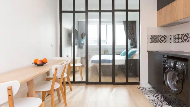 A modern kitchen with a washing machine, a dining area with a table and chairs, and a sliding glass door leading to a bedroom. - Modern apartment with plenty of public transport! (Neuilly-sur-Seine)