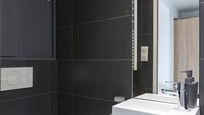 A modern bathroom with dark tiles, a white toilet, a wooden vanity, and a mirror. - Modern apartment with plenty of public transport! (Neuilly-sur-Seine)