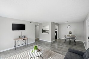Ein modernes Wohnzimmer mit einem Flachbildfernseher, einem kleinen Tisch mit Pflanze und einem grauen Sofa.
