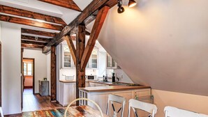 Un comedor con una mesa de madera rústica, sillas blancas y una cesta tejida. Se ve una cocina con armarios blancos y un microondas al fondo.