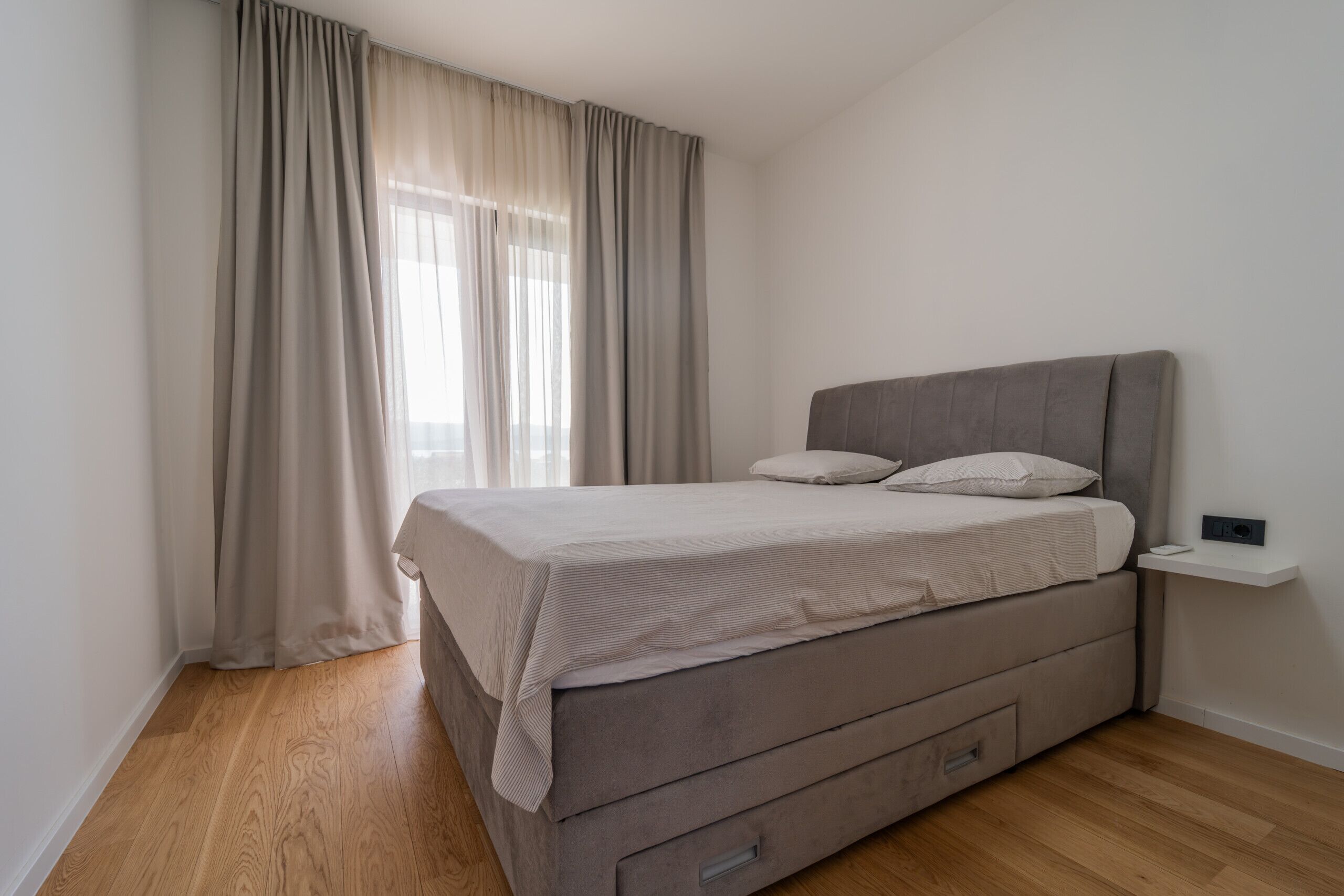 A bedroom with a bed, wooden flooring, a window with curtains, and a bedside table.