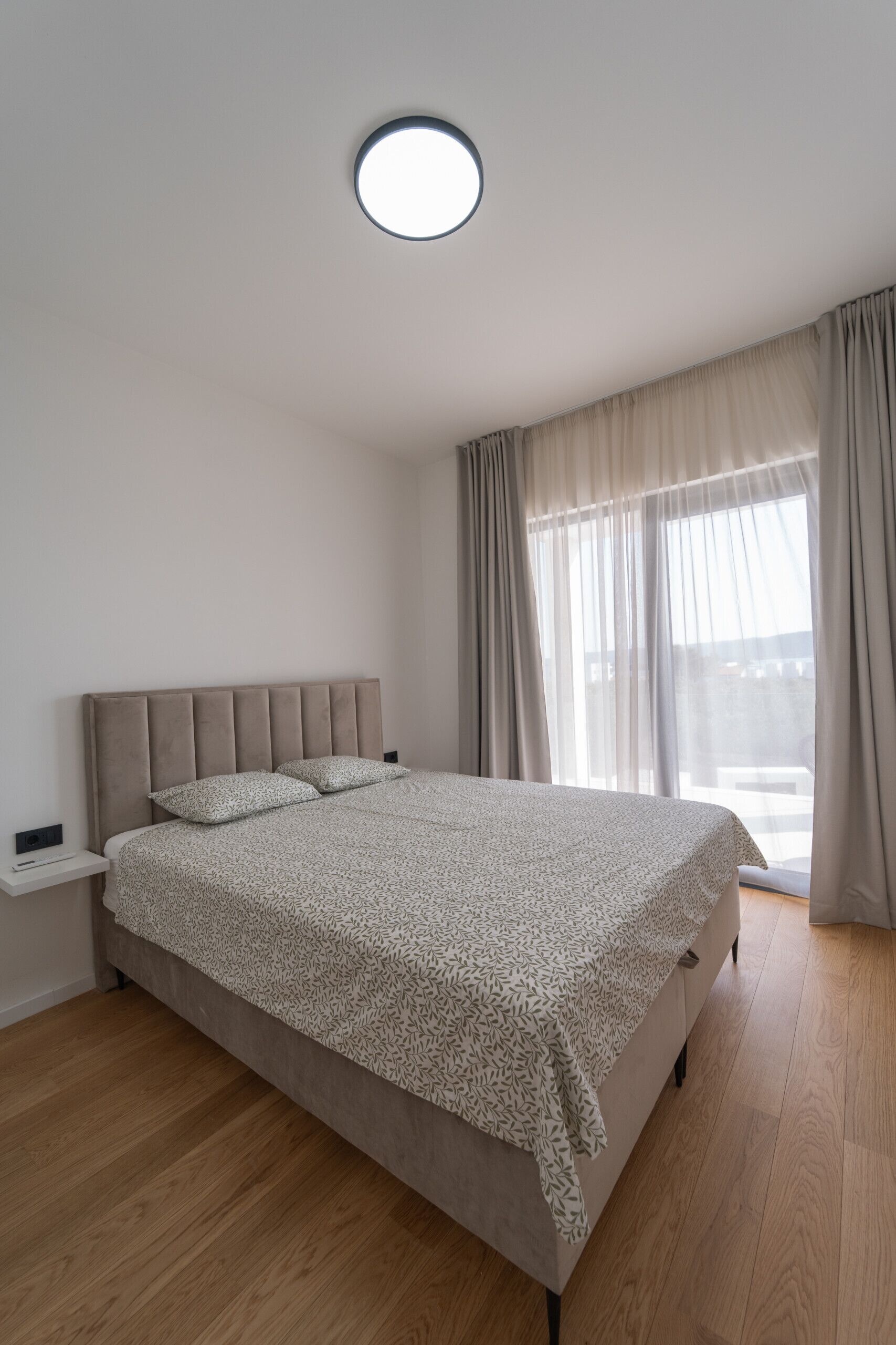 A modern bedroom with a large bed, a bedside table, and a view of the outdoors through sheer curtains.