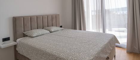 A modern bedroom with a large bed, a bedside table, and a view of the outdoors through sheer curtains.