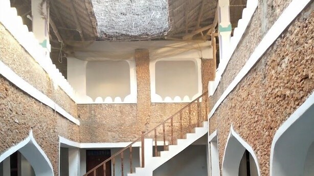 Um interior com vários níveis, escadaria central, paredes de pedra e janelas em arco.