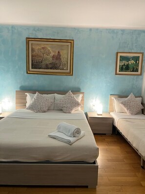 Una cama doble con una colcha blanca y dos almohadas, una mesita de noche con una lámpara, y dos cuadros enmarcados en la pared.