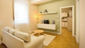 A living room with a sofa, a small round table, a TV on a white shelf, and a kitchen in the background. - CASA AL GELSO, authentic Tuscan atmosphere (Lucca)