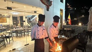 Deux chefs préparent des plats en plein air, de nuit, avec un restaurant en arrière-plan.